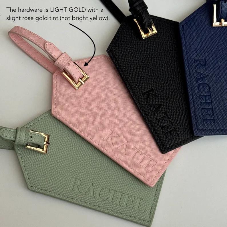 May include: A collection of luggage tags in various colors: pink, black, navy blue, and green. Each tag features a name embossed in capital letters. The hardware is light gold with a slight rose gold tint.