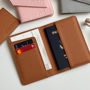 May include: A brown leather passport holder with a passport and boarding pass inside. The holder has slots for credit cards. Other personalized passport holders in pink, gray, and brown are visible, with names embossed on them.