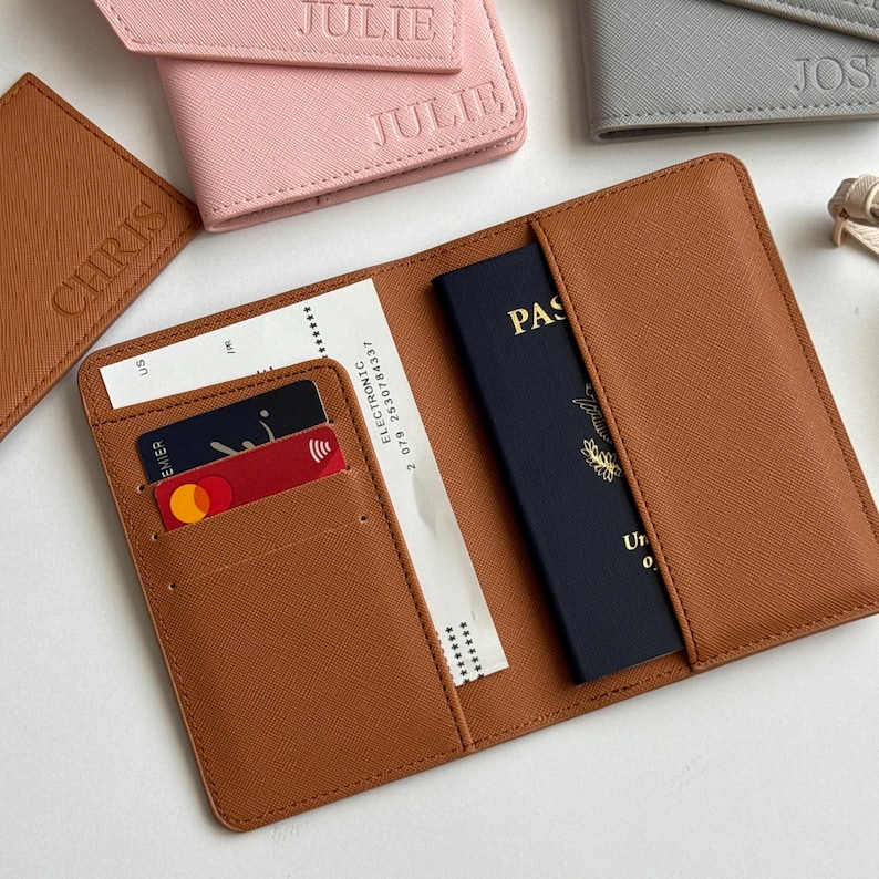 May include: A brown leather passport holder with a passport and boarding pass inside. The holder has slots for credit cards. Other personalized passport holders in pink, gray, and brown are visible, with names embossed on them.