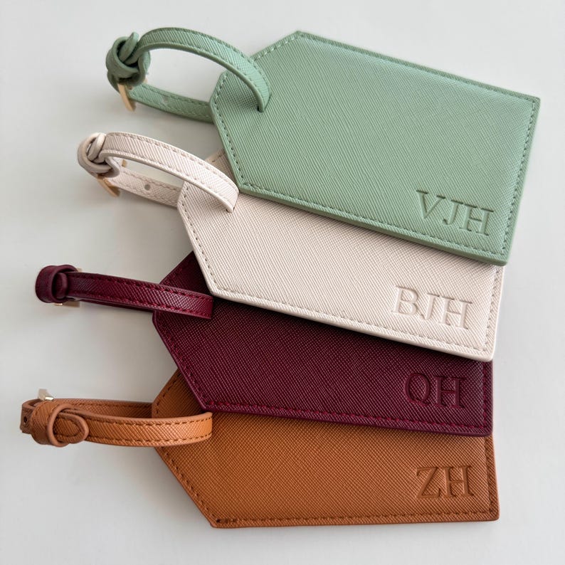 May include: Four rectangular luggage tags with pointed ends, each featuring a textured surface and a matching buckled strap. The tags are colored sage green, cream, burgundy, and tan, and each is embossed with unique three-letter initials.