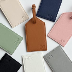 May include: A collection of personalized luggage tags in various colors, including brown, navy, pink, and green. Each tag features embossed initials, with a leather strap for attachment. The tags are arranged on a white surface.