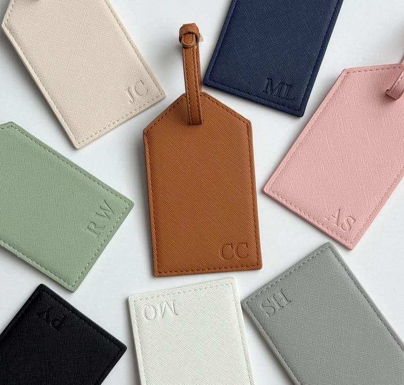 May include: A collection of personalized luggage tags in various colors, including brown, navy, pink, and green. Each tag features embossed initials, with a leather strap for attachment. The tags are arranged on a white surface.
