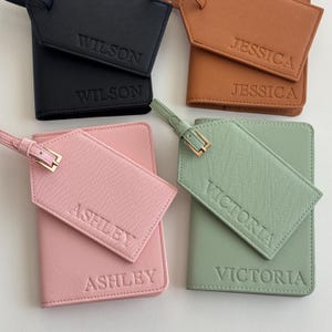 May include: Four sets of passport holders and matching luggage tags in black, tan, pink, and sage green. Each item features a textured finish and is embossed with names: Wilson, Jessica, Ashley, and Victoria. Each tag has a gold-tone buckle.