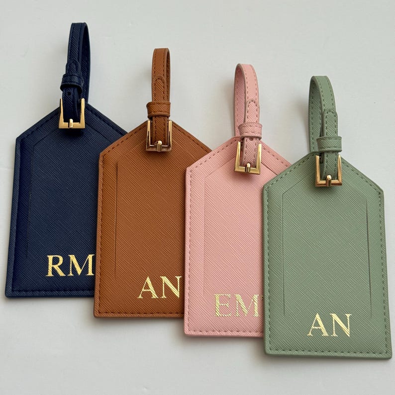 Puede incluir: Cuatro etiquetas de equipaje personalizadas en azul marino, marr&oacute;n, rosa y verde salvia. Cada etiqueta tiene una hebilla y una correa doradas, con iniciales doradas. Las etiquetas son rectangulares con una parte superior en forma de casa.