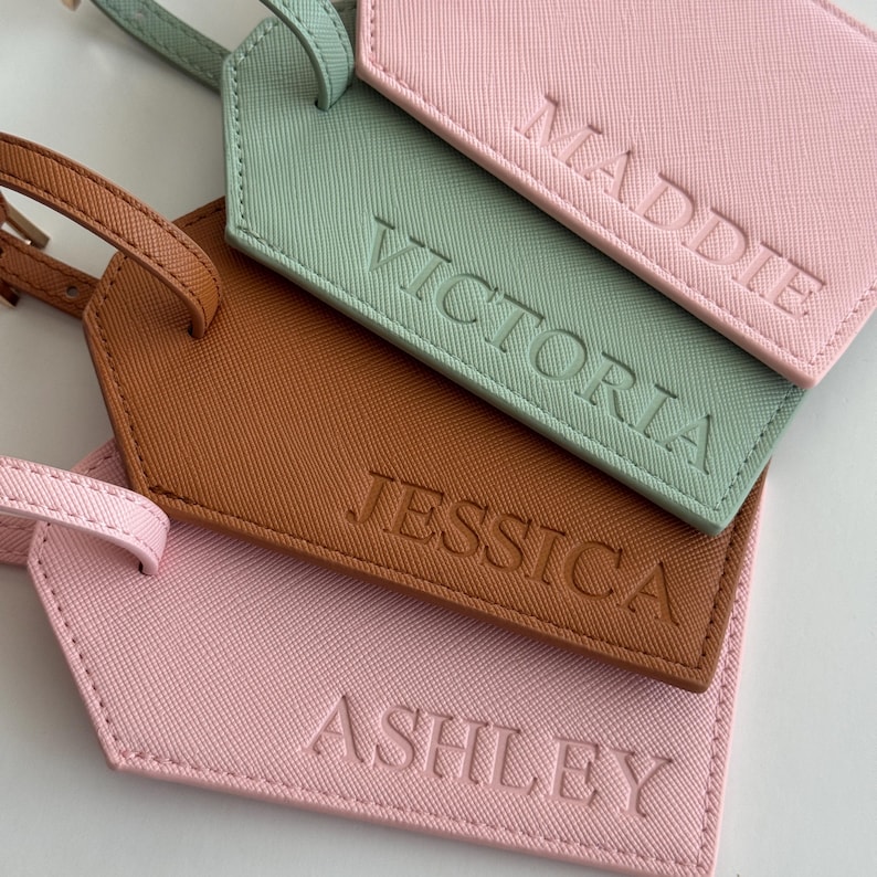 May include: Four rectangular luggage tags with straps, featuring a textured finish. The tags are colored light pink, mint green, and tan. Each tag is embossed with a name in capital letters: MADDIE, VICTORIA, JESSICA, and ASHLEY, respectively.