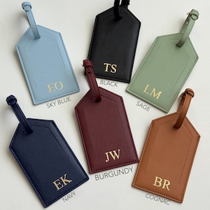 Puede incluir: Un conjunto de etiquetas de equipaje personalizadas en varios colores: azul cielo, negro, verde salvia, azul marino, burdeos y co&ntilde;ac. Cada etiqueta presenta iniciales doradas y el nombre del color. Forma rectangular con correa.