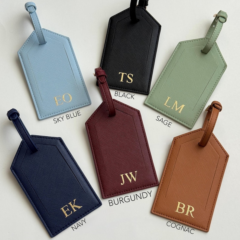 Puede incluir: Un conjunto de etiquetas de equipaje personalizadas en varios colores: azul cielo, negro, verde salvia, azul marino, burdeos y co&ntilde;ac. Cada etiqueta presenta iniciales doradas y el nombre del color. Forma rectangular con correa.