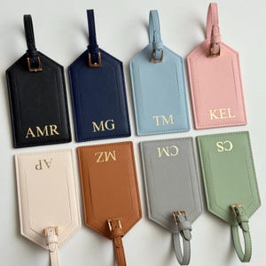 Puede incluir: Una colecci&oacute;n de etiquetas de equipaje personalizadas en varios colores, incluyendo negro, azul marino, azul claro, rosa, beige, marr&oacute;n, gris y verde. Cada etiqueta presenta letras doradas con iniciales y una correa a juego con una hebilla dorada.