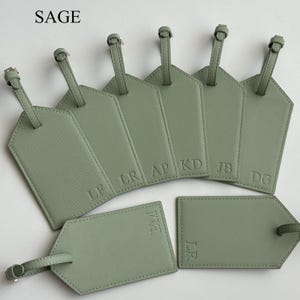 May include: Sage green luggage tags with embossed initials. The tags have a textured finish and a strap with a buckle. The word "SAGE" is in the top left corner.