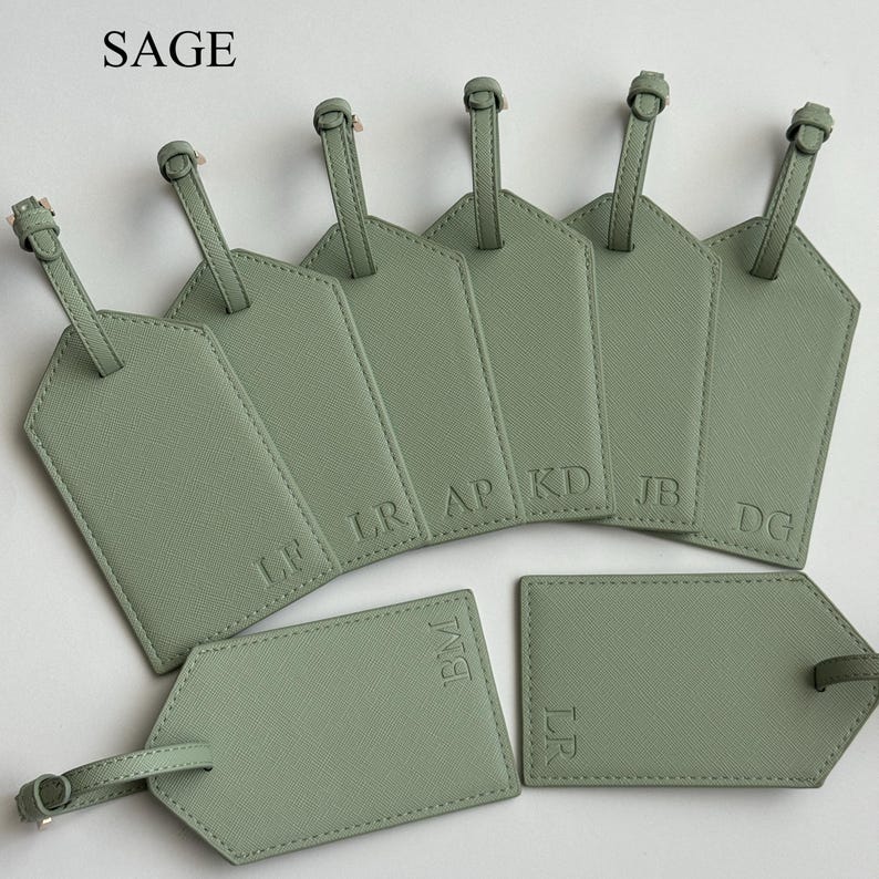 May include: Sage green luggage tags with embossed initials. The tags have a textured finish and a strap with a buckle. The word "SAGE" is in the top left corner.