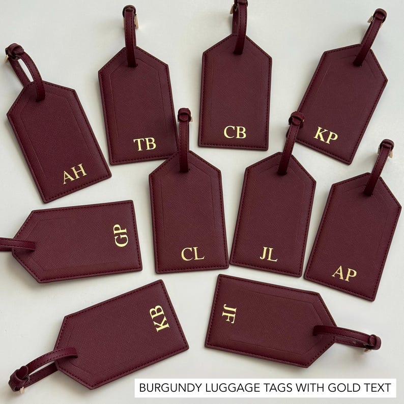 Puede incluir: Etiquetas de equipaje burdeos con texto dorado. Las etiquetas son rectangulares con una parte inferior puntiaguda y una correa en la parte superior. Cada etiqueta tiene iniciales doradas. El texto "BURGUNDY LUGGAGE TAGS WITH GOLD TEXT" est&aacute; en la parte inferior.