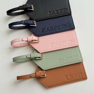 May include: A collection of personalized luggage tags in various colors: black, navy, pink, sage green, and brown. Each tag features a name embossed in a matching color, with a strap and buckle for attachment. The names are Jared, Caroline, Caitlin, Emily, and Leah.