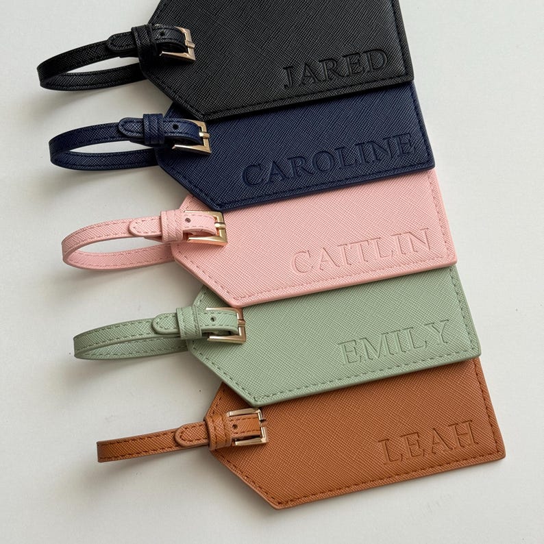May include: A collection of personalized luggage tags in various colors: black, navy, pink, sage green, and brown. Each tag features a name embossed in a matching color, with a strap and buckle for attachment. The names are Jared, Caroline, Caitlin, Emily, and Leah.
