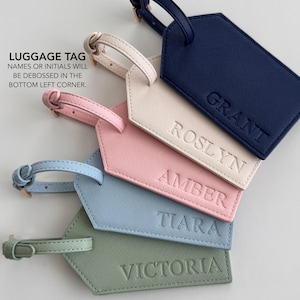 May include: Five rectangular luggage tags with buckled straps, each featuring a textured surface and a debossed name. The colors include navy blue with GRANT, cream with ROSLYN, pink with AMBER, light blue with TIARA, and sage green with VICTORIA.