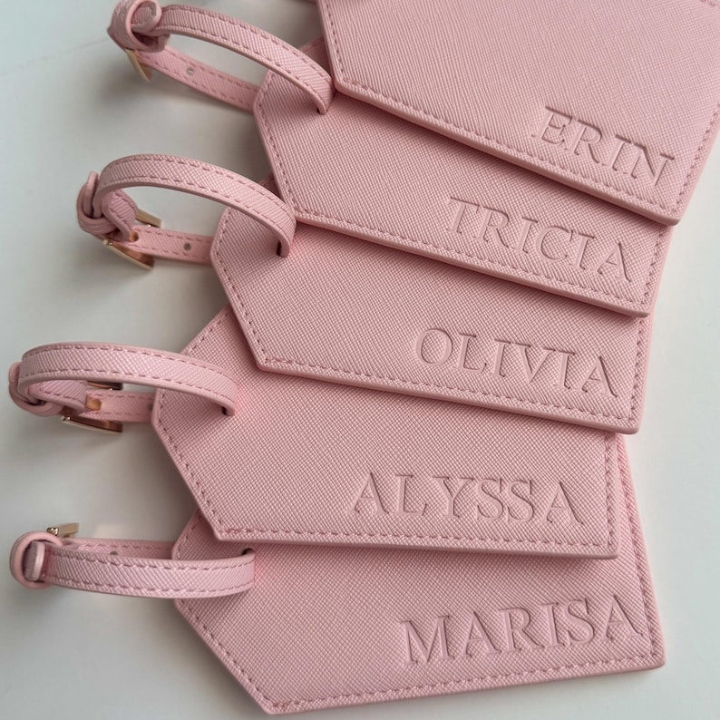 May include: Pink luggage tags with gold-colored buckles and straps. Each tag has a name embossed on it: ERIN, TRICIA, OLIVIA, ALYSSA, and MARISA. The tags are made of a textured material.