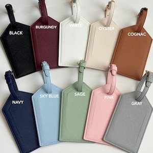 May include: A collection of luggage tags in various colors, including black, burgundy, white, oyster, cognac, navy, sky blue, sage, pink, and gray. Each tag has a strap and a gold-colored buckle, with the color name printed on each tag.