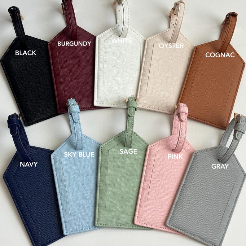 May include: A collection of luggage tags in various colors, including black, burgundy, white, oyster, cognac, navy, sky blue, sage, pink, and gray. Each tag has a strap and a gold-colored buckle, with the color name printed on each tag.