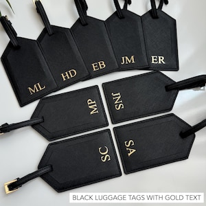 Puede incluir: Etiquetas de equipaje negras con letras doradas, con iniciales como ML, HD y EB. Cada etiqueta tiene una correa y una hebilla. El texto "BLACK LUGGAGE TAGS WITH GOLD TEXT" est&aacute; en la parte inferior.
