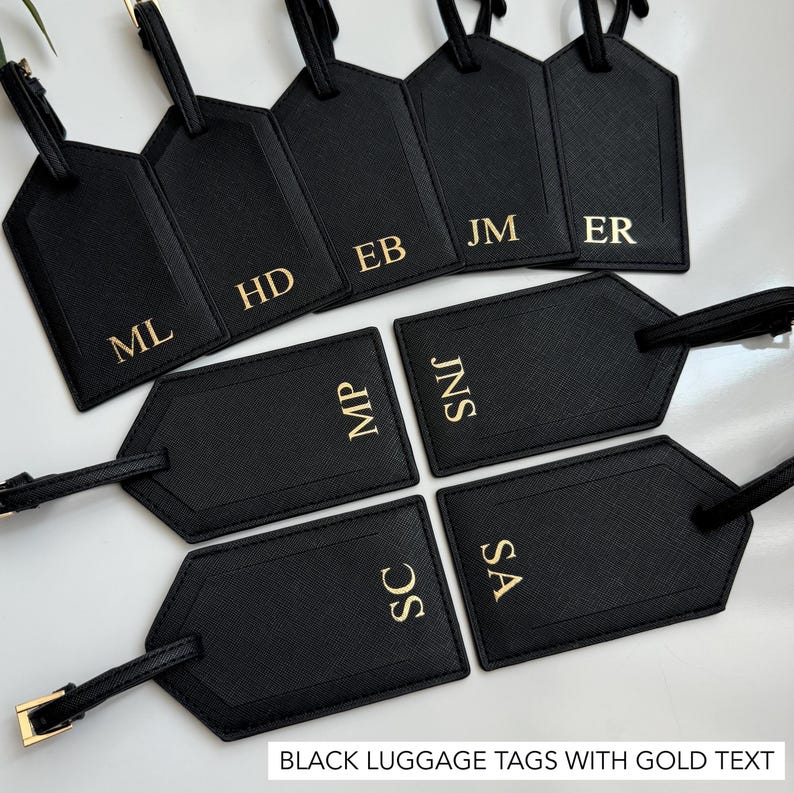 Puede incluir: Etiquetas de equipaje negras con letras doradas, con iniciales como ML, HD y EB. Cada etiqueta tiene una correa y una hebilla. El texto "BLACK LUGGAGE TAGS WITH GOLD TEXT" est&aacute; en la parte inferior.