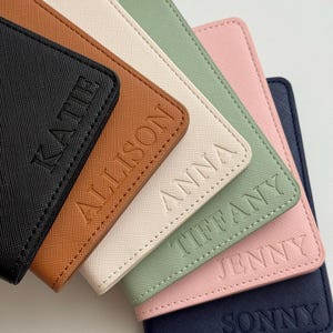 May include: A collection of personalized passport holders in various colors, including black, brown, cream, green, pink, and navy. Each holder is embossed with a name: Kate, Allison, Anna, Tiffany, Jenny, and Sonny.