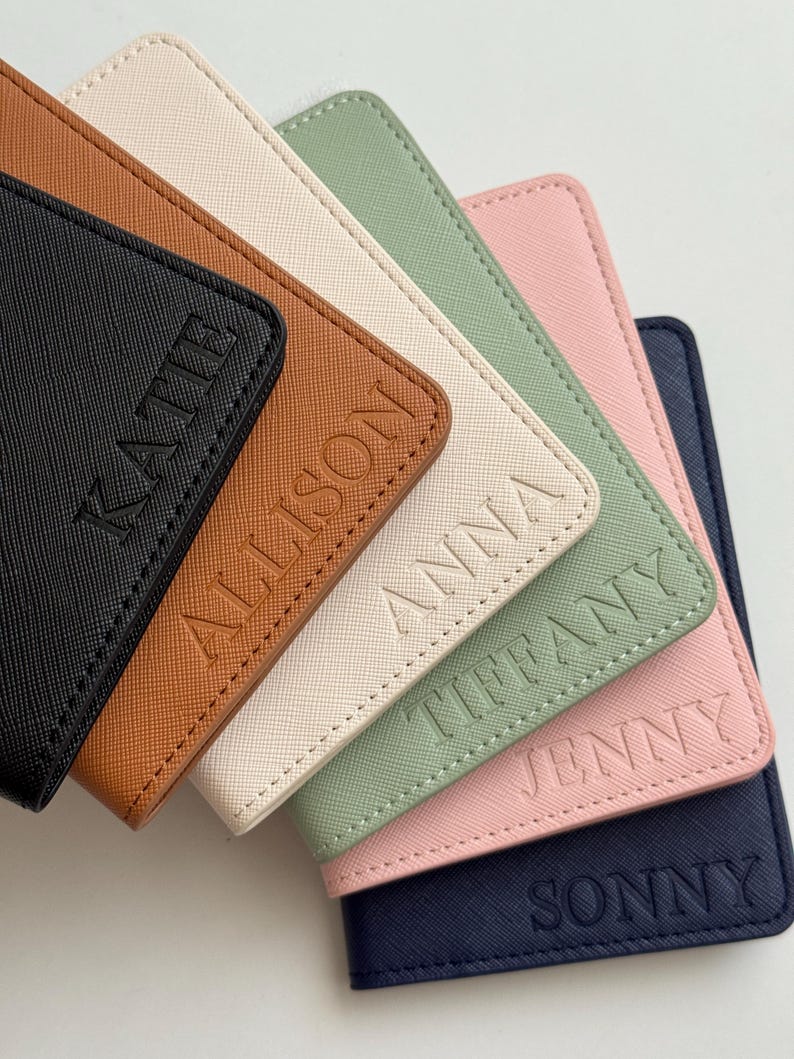 May include: A collection of personalized passport holders in various colors, including black, brown, cream, green, pink, and navy. Each holder is embossed with a name: Kate, Allison, Anna, Tiffany, Jenny, and Sonny.
