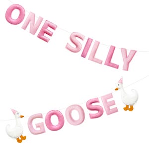 May include: Pink felt banner with the words "ONE SILLY GOOSE". The banner includes two white geese wearing party hats. The letters are in varying shades of pink and are strung on a white string.