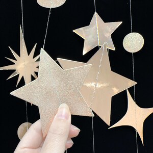 May include: A set of gold glitter star and circle shaped paper decorations hanging from strings. The stars are different sizes and have a shimmery finish.