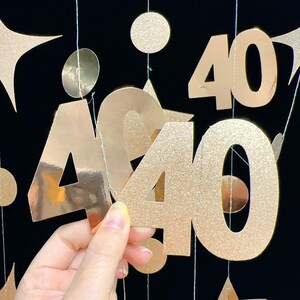 May include: Gold glitter paper cutouts of the number 40, hanging from strings. The cutouts are in various sizes and shapes, including circles and stars. The background is black.