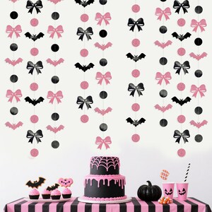 May include: A Halloween-themed party scene with pink and black decorations. Pink and black hanging garlands with bows, bats, and circles. A black cake with pink icing, cupcakes, and a pumpkin on a striped table.