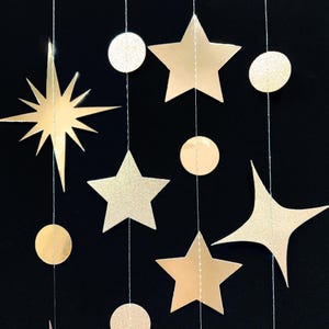 May include: A gold garland with stars and circles hanging from a black background. The garland is made of paper and has a sparkly finish.