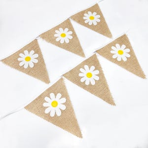 May include: A burlap banner with white daisy flowers with yellow centers. The banner is strung together with white ribbon.