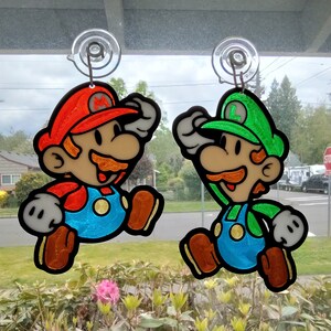 Puede incluir: Dos personajes de dibujos animados coloridos, Mario y Luigi, est&aacute;n colgados de ventosas en una ventana. Mario lleva un sombrero rojo y un mono, mientras que Luigi lleva un sombrero verde y un mono. Ambos sonr&iacute;en y tienen un estilo de dibujos animados.