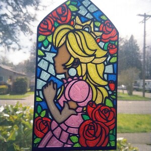 Op de afbeelding: Een hangend ornament van glas-in-lood met een prinses met blond haar en een roze jurk, omringd door rode rozen. De achtergrond is een moza&iuml;ek van blauw, groen en geel glas.
