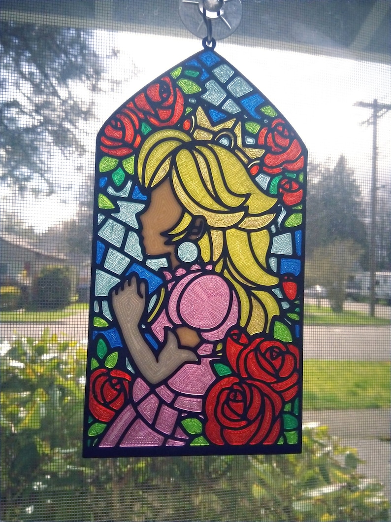 Op de afbeelding: Een hangend ornament van glas-in-lood met een prinses met blond haar en een roze jurk, omringd door rode rozen. De achtergrond is een moza&iuml;ek van blauw, groen en geel glas.