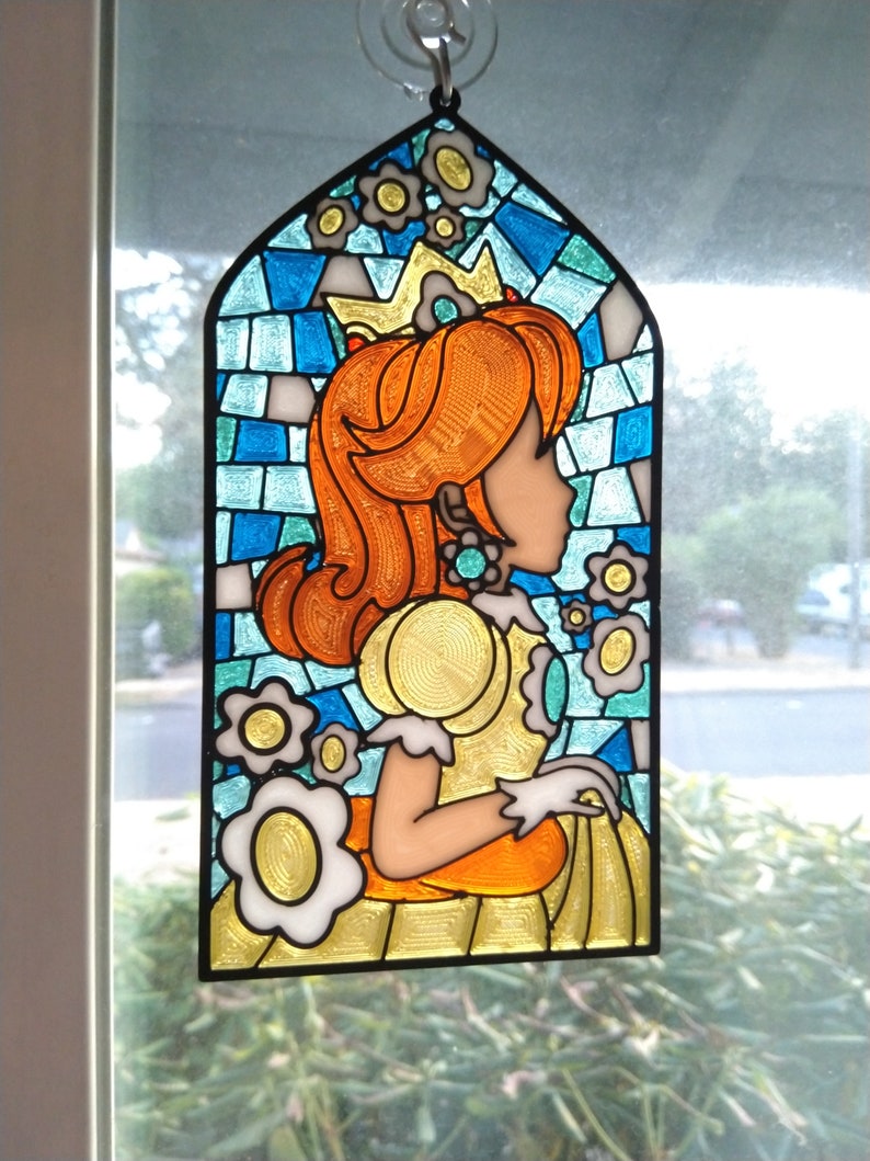 May include: A stained glass window hanging ornament featuring Princess Peach from the Super Mario Bros. video game series. The ornament is in a blue, yellow, and orange color scheme and features a crown, flowers, and a detailed portrait of Princess Peach.