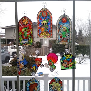 Op de afbeelding: Een verzameling van gebrandschilderd glas raamhangers met personages uit populaire videogames. De personages zijn Link uit The Legend of Zelda, Mario en Luigi uit Super Mario Bros., Sonic the Hedgehog, Donkey Kong en Princess Peach. De hangers zijn kleurrijk en gedetailleerd, met ingewikkelde ontwerpen en levendige kleuren.