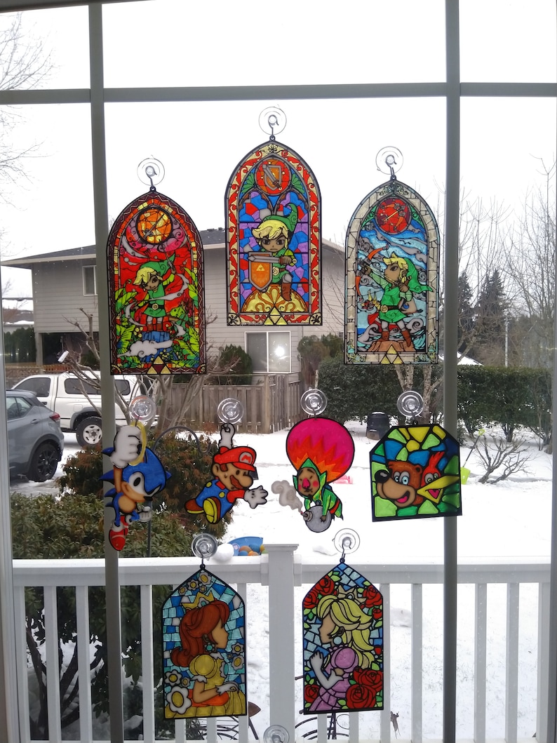 Op de afbeelding: Een verzameling van gebrandschilderd glas raamhangers met personages uit populaire videogames. De personages zijn Link uit The Legend of Zelda, Mario en Luigi uit Super Mario Bros., Sonic the Hedgehog, Donkey Kong en Princess Peach. De hangers zijn kleurrijk en gedetailleerd, met ingewikkelde ontwerpen en levendige kleuren.
