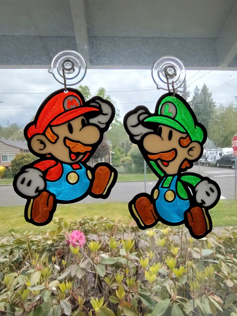 Puede incluir: Dos personajes de dibujos animados coloridos, Mario y Luigi, est&aacute;n colgados de ventosas en una ventana. Mario lleva un sombrero rojo y un mono, mientras que Luigi lleva un sombrero verde y un mono. Ambos sonr&iacute;en y tienen un estilo de dibujos animados.