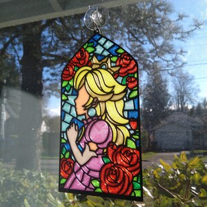 Op de afbeelding: Een gebrandschilderd raamdecoratie met een cartoonprinses met blond haar en een roze jurk. De prinses wordt omringd door rode rozen en groene bladeren. De decoratie is gemaakt van helder plastic met een zwarte rand.