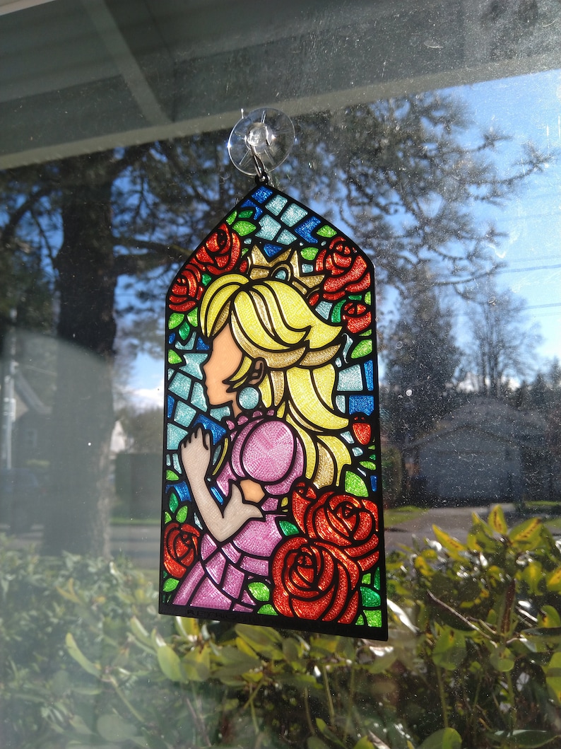 Op de afbeelding: Een gebrandschilderd raamdecoratie met een cartoonprinses met blond haar en een roze jurk. De prinses wordt omringd door rode rozen en groene bladeren. De decoratie is gemaakt van helder plastic met een zwarte rand.