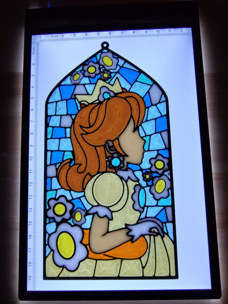 May include: A stained glass window featuring a princess with a crown and a yellow dress. The princess is surrounded by blue and white flowers. The window is framed in black and is approximately 19 inches tall.