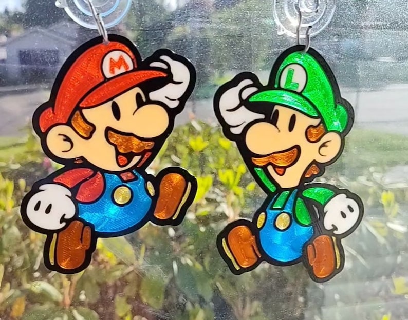 Puede incluir: Dos pendientes de personajes de papel, uno rojo y otro verde. El personaje rojo tiene un sombrero rojo con una 'M' y un bigote marr&oacute;n. El personaje verde tiene un sombrero verde con una 'L' y un bigote marr&oacute;n.