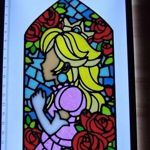 Op de afbeelding: Een glas-in-loodraamontwerp met een prinses met blond haar en een roze jurk. De prinses wordt omringd door rode rozen en groene bladeren. Het ontwerp is omlijst met zwart.