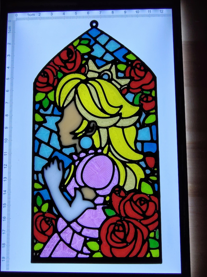 Op de afbeelding: Een glas-in-loodraamontwerp met een prinses met blond haar en een roze jurk. De prinses wordt omringd door rode rozen en groene bladeren. Het ontwerp is omlijst met zwart.