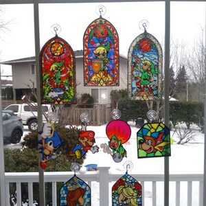 May include: A collection of stained glass window hangings featuring characters from popular video games. The characters include Link from The Legend of Zelda, Mario and Luigi from Super Mario Bros., Sonic the Hedgehog, Donkey Kong, and Princess Peach. The hangings are colorful and detailed, with intricate designs and vibrant colors.