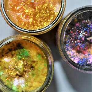 May include: Three glass jars with lids, each filled with a different colored wax and glitter. The first jar has gold glitter and orange wax, the second jar has green and gold glitter with yellow wax, and the third jar has pink, blue, and purple glitter with a dark purple wax.