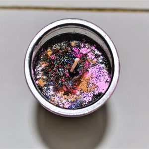 May include: A black candle with a silver container and a wooden wick. The candle is decorated with pink, gold, and blue glitter.