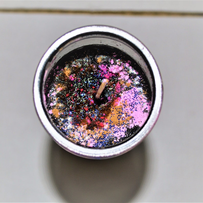 May include: A black candle with a silver container and a wooden wick. The candle is decorated with pink, gold, and blue glitter.