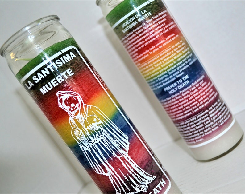 May include: Two glass candle jars with rainbow colored wax and white labels. The label on the left jar reads "La Santisima Muerte" and the label on the right jar reads "Oracion de la Santisima Muerte" and "Prayer to the Holy Death".