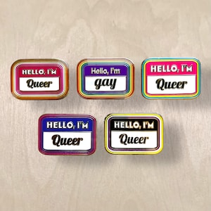 May include: Five rectangular enamel pins with rounded corners. The pins have a gold border and feature the phrases "Hello, I'm Queer" and "Hello, I'm gay" in various color schemes, including pink, purple, and blue.